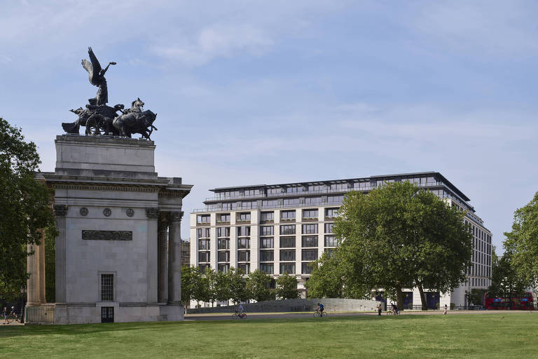 A imagem mostra um monumento com uma escultura no topo， representando uma figura alada em um carro puxado por cavalos. O monumento é de pedra clara e possui colunas. Ao fundo， há um edifício moderno de vários andares com janelas grandes e uma fachada clara. O espaço ao redor é um gramado verde， onde algumas pessoas estão andando de bicicleta. O céu está claro com algumas nuvens.