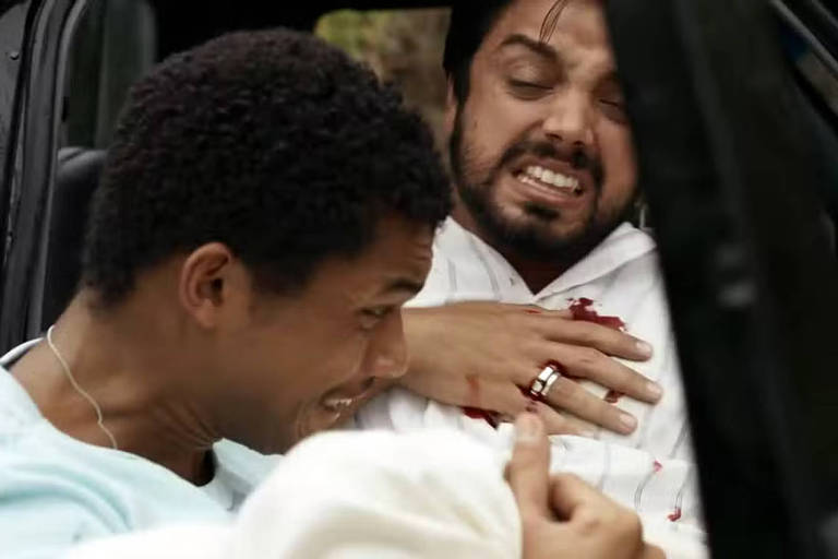 A imagem mostra duas pessoas dentro de um carro. Um homem com cabelo crespo e pele clara está segurando um homem com cabelo escuro e pele morena, que parece estar ferido, com sangue visível em sua camisa branca. O homem ferido está com uma expressão de dor, enquanto o outro parece angustiado. O ambiente é interno de um carro, com janelas e um fundo desfocado.