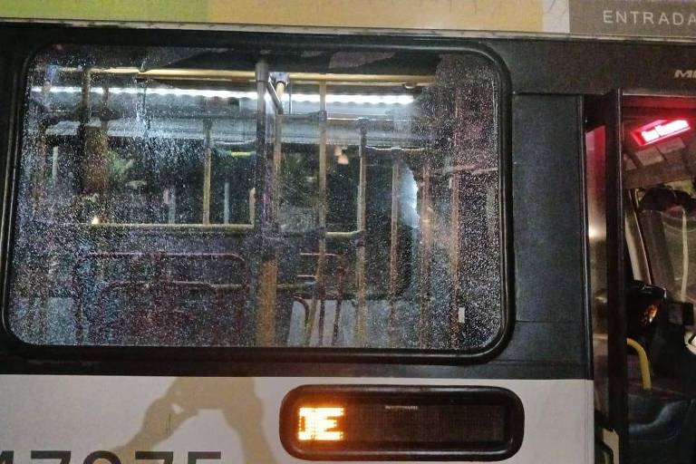 A imagem mostra o interior de um ônibus， com janelas embaçadas e iluminação interna. É possível ver o reflexo de pessoas e objetos do lado de fora. A parte externa do ônibus é visível， com um número de identificação na lateral.