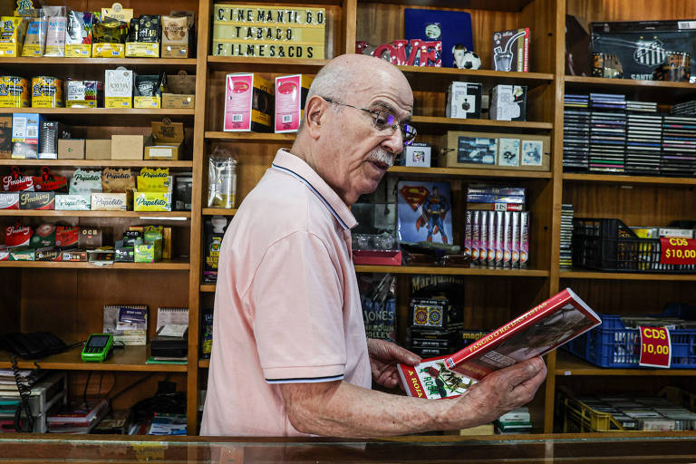 Um homem idoso, com óculos e cabelo grisalho, está em pé atrás de um balcão de loja, segurando um livro. Ele usa uma camisa polo rosa e parece estar lendo ou examinando o conteúdo do livro. Ao fundo, há prateleiras repletas de produtos variados, incluindo caixas de cigarros, CDs e outros itens. A loja tem um ambiente acolhedor e está bem organizada.