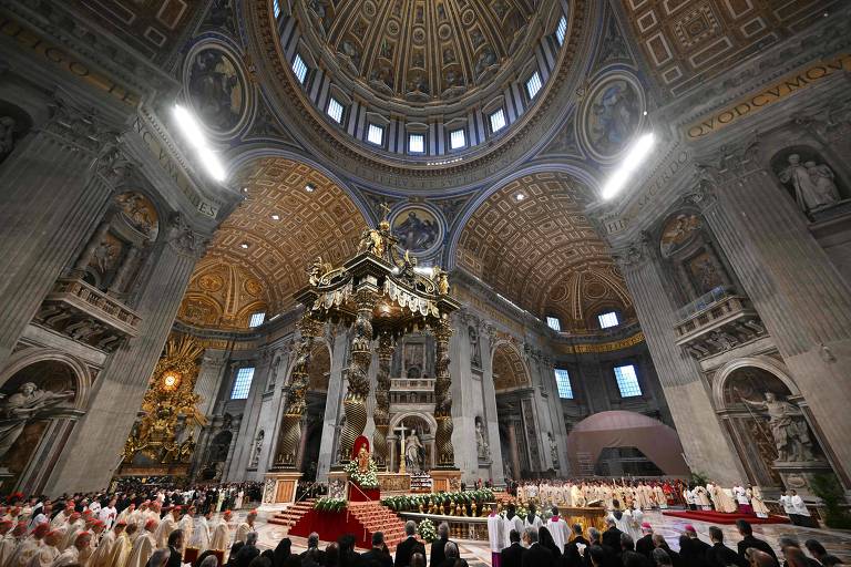 A imagem mostra o interior da Basílica de São Pedro， com uma grande cúpula ornamentada e alta， iluminada por janelas. No centro， há um altar decorado e um grande candelabro pendurado. Várias pessoas estão reunidas em torno do altar， algumas vestindo vestes litúrgicas. O ambiente é grandioso， com colunas e detalhes arquitetônicos elaborados.