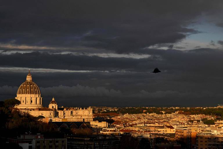 A imagem mostra uma vista do Vaticano ao entardecer， com nuvens escuras no céu. A cúpula da Basílica de São Pedro é visível à esquerda， iluminada pela luz do sol que se põe. A cidade de Roma se estende ao fundo， com uma tonalidade dourada devido à luz do crepúsculo. Um objeto escuro está presente no céu， mas não é possível identificar o que é.