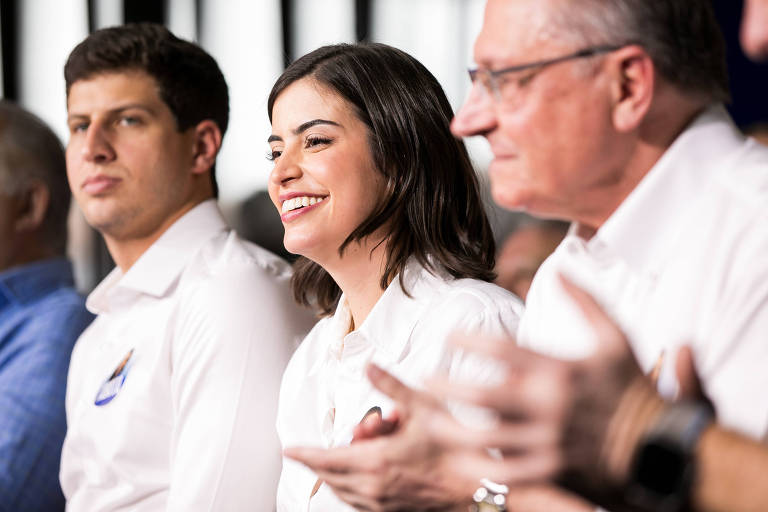 A imagem mostra um grupo de três pessoas sentadas em um evento. À esquerda， o prefeito de Recife， João Campos， com cabelo curto e camisa branca. No centro， Tabata Amaral sorridente com cabelo escuro e também vestindo uma camisa branca. À direita， o vice-presidente Geraldo Alckmincom óculos e camisa branca， que parece estar aplaudindo. O fundo é desfocado， sugerindo um ambiente de evento.