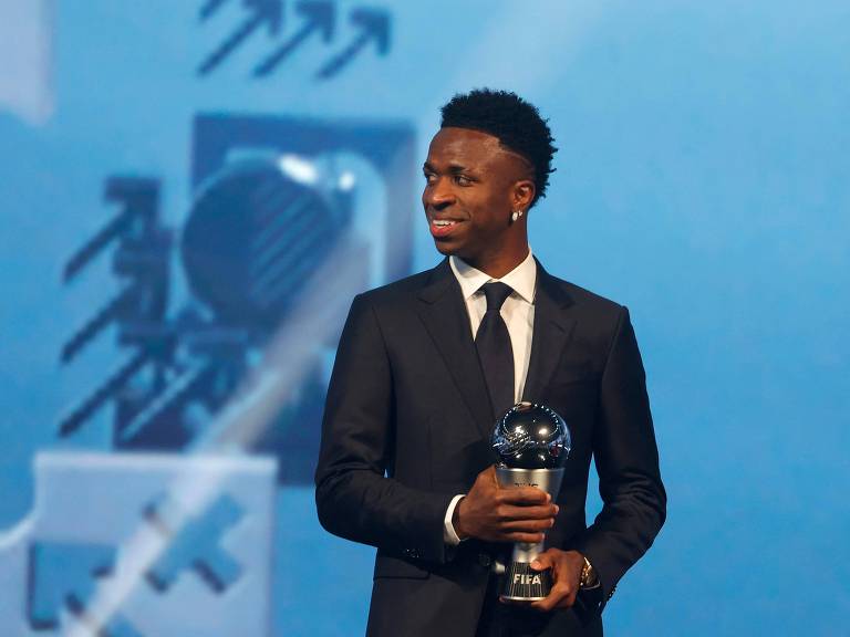 Vestido com terno e gravata pretos e camisa branca， Vinicius Junior， durante cerimônia de premiação em Doha (Qatar)， olha para o seu lado direito segurando o troféu The Best， que ele conquistou por ter sido eleito pela Fifa o melhor jogador do mundo na temporada 2023/2024