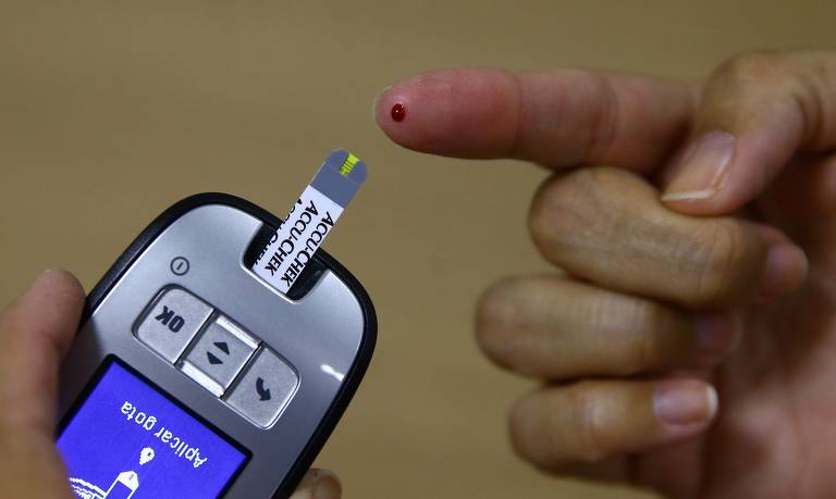 Foto mostra mão de uma pessoa branca com um pouco de sangue no dedo indicador e uma aparelho medidor de insulina ao lado.