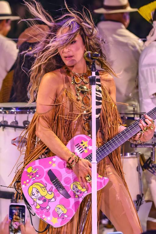 Uma artista feminina está se apresentando em um palco, segurando uma guitarra rosa decorada com desenhos de personagens. Ela usa um traje com franjas e acessórios brilhantes. O cabelo dela está esvoaçando, e ao fundo, há músicos com instrumentos, incluindo tambores. A atmosfera é vibrante e festiva.