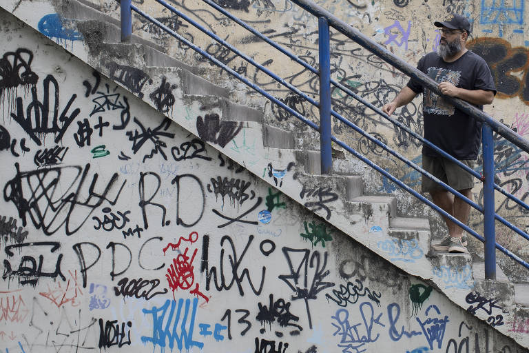 Um homem negro de barba, óculos e boné está parado em uma escada. As paredes ao redor estão cobertas de grafites coloridos e desenhos. O homem usa uma camiseta escura e calças claras, e observa o ambiente enquanto se apoia na grade azul da escada.