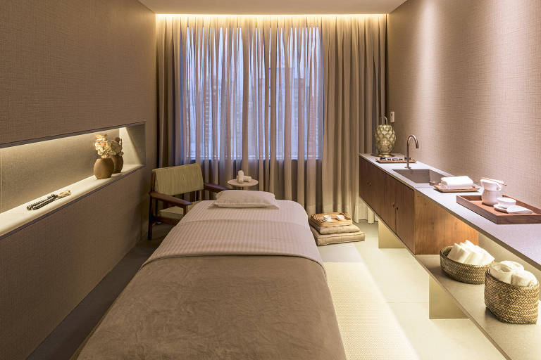 A imagem mostra um ambiente de spa com uma cama de massagem coberta por um lençol claro. Ao fundo, há uma janela com cortinas longas e suaves. À esquerda, uma prateleira iluminada exibe um vaso com flores e um colar. Um sofá verde está ao lado da cama, e uma mesa pequena está próxima. À direita, uma bancada de madeira com uma pia e utensílios de spa, incluindo toalhas e um recipiente, está visível. O ambiente é iluminado de forma suave, criando uma atmosfera relaxante.