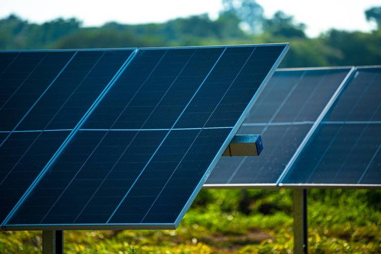 A imagem mostra painéis solares dispostos em um campo. Os painéis são retangulares e possuem uma superfície escura e brilhante， refletindo a luz do sol. Ao fundo， há uma vegetação verde e um céu claro.