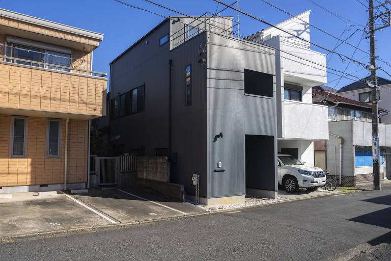 A imagem mostra duas casas em uma área urbana. À esquerda, uma casa de cor clara com uma varanda. À direita, uma casa moderna de cor cinza escuro, com janelas retangulares e uma entrada para garagem. No fundo, há uma casa branca e um carro estacionado na calçada. O céu está limpo e azul.