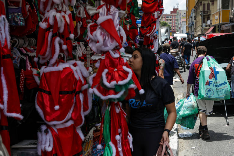 A imagem mostra uma rua movimentada com várias fantasias de Natal expostas em um comércio. À esquerda, há várias roupas vermelhas com detalhes brancos, típicas do Natal, como chapéus e trajes. Uma mulher com uma camiseta preta está olhando para as roupas. Ao fundo, outras pessoas caminham pela rua, algumas carregando sacolas de compras.