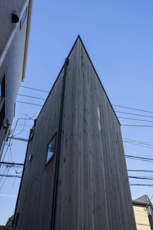 A imagem mostra uma casa com um design angular, vista de baixo para cima. A estrutura é feita de madeira e tem um telhado pontudo. Ao fundo, é possível ver fios elétricos e um céu azul claro. A casa está situada entre outras edificações.