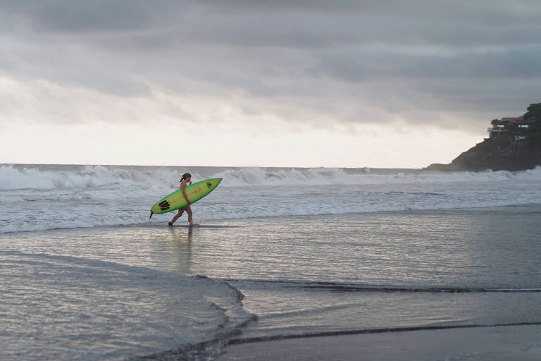 Uma surfista caminha pela praia， segurando uma prancha de surf verde. O mar está agitado， com ondas quebrando ao fundo. O céu está nublado， criando uma atmosfera dramática. À direita， há uma colina com algumas construções visíveis.