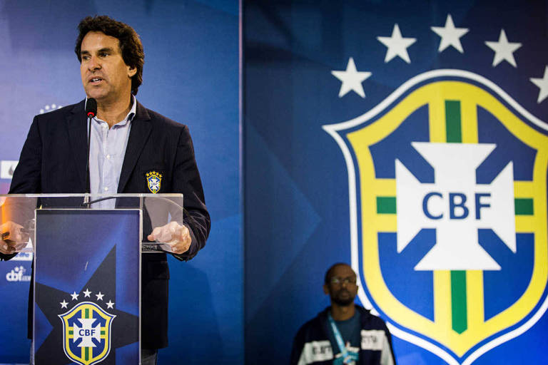 Um homem está falando em um microfone em uma coletiva de imprensa， com um fundo que exibe o escudo da Confederação Brasileira de Futebol (CBF). Ele usa um paletó escuro e uma camisa clara. Ao fundo， há um homem em pé， aparentemente assistindo à coletiva.