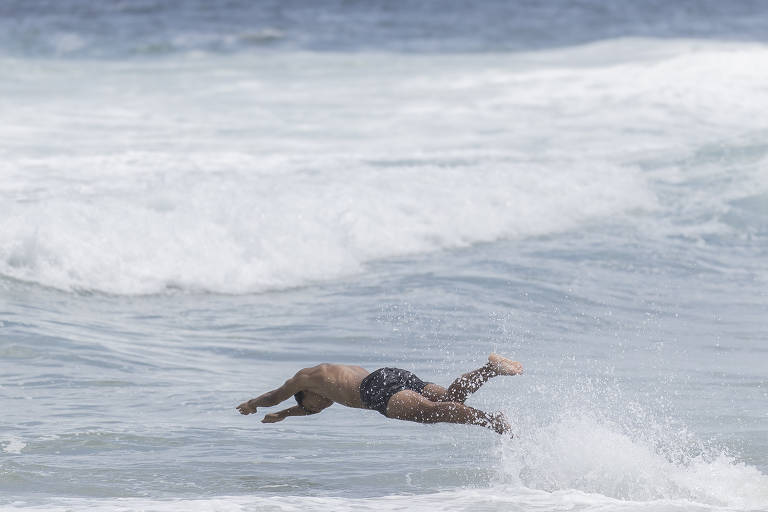 Um homem está mergulhando nas ondas do mar, com o corpo inclinado para frente e as pernas estendidas. O mar está agitado, com ondas brancas e espumantes ao fundo. O céu é claro e a água parece azul.