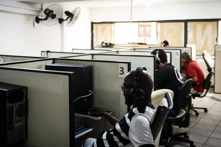 A imagem mostra um ambiente de trabalho com várias estações de computador. Há três pessoas visíveis, cada uma usando fones de ouvido e concentradas em suas telas. As estações são divididas por painéis, e o espaço é iluminado por luz natural que entra pelas janelas. Ventiladores de teto estão visíveis no teto.