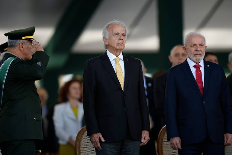 Várias pessoas enfileiradas diante de cadeiras. Na frente, dois homens brancos, Múcio, um homen de cabelos brancos, vestido de terno preto e gravata amarela, e Lula, um homem de cabelos e barba branca, vestido de terno escuro e uma gravata vermelha. No canto, um homem branco com a farda do Exército faz uma saudação militar, posicionando a mão na direção da testa