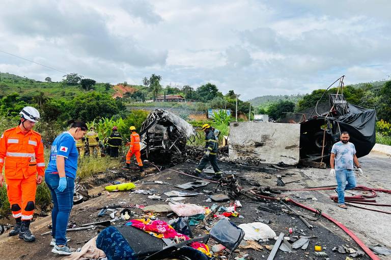 A imagem mostra o local de um acidente de trânsito, onde um caminhão está tombado na estrada. Há destroços espalhados pelo chão, incluindo roupas e objetos pessoais. Várias pessoas, incluindo socorristas em uniformes laranja e azul, estão presentes na cena, algumas examinando os destroços e outras ajudando. O céu está nublado e a vegetação é visível ao fundo.