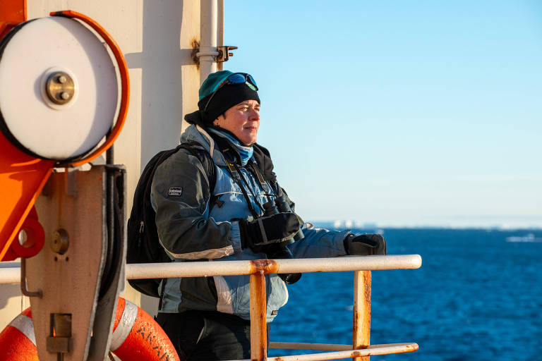 Uma pessoa está em um barco， apoiada em uma grade， olhando para o horizonte. Ela usa uma jaqueta azul e preta， um gorro e óculos de sol. Ao fundo， há água e um céu claro. Um salva-vidas laranja está visível à esquerda.