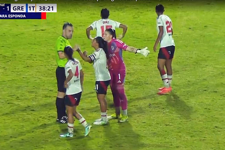 A imagem mostra um momento de discussão em um jogo de futebol. Uma jogadora da equipe com uniforme branco e vermelho está conversando com o árbitro， enquanto outras jogadoras da mesma equipe estão ao fundo. A goleira， vestida de rosa， também está envolvida na situação. O campo está coberto de grama e a iluminação é adequada para a partida.
