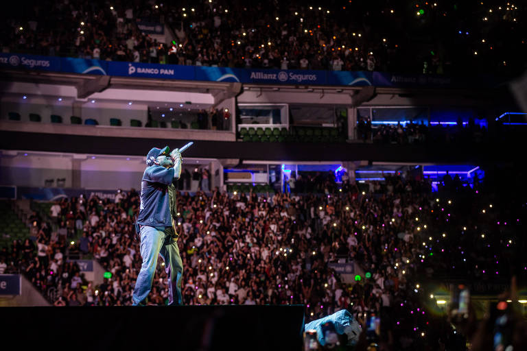 Cantor é visto em cima do palco durante performance; público e estrutura do estádio são vistos ao fundo, com luzes de celular.