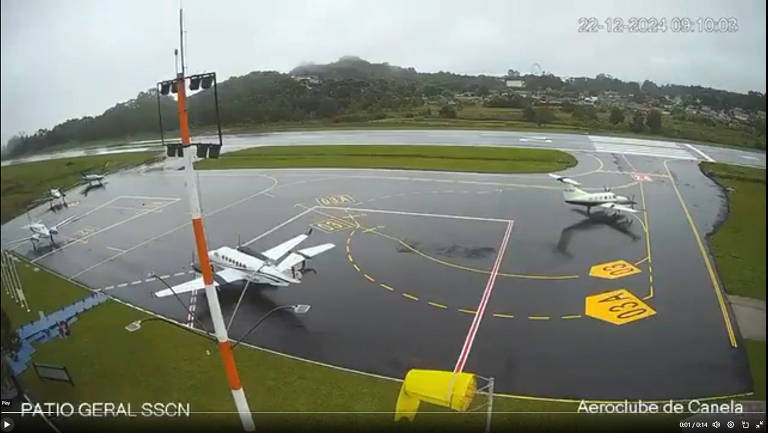 Imagem do pátio do Aeródromo de Canela em um dia chuvoso. Vários aviões pequenos estão estacionados na pista molhada, com marcas de sinalização visíveis no chão. O céu está nublado e há uma vegetação ao fundo.