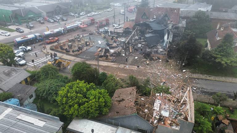 A imagem mostra uma cena de destruição em uma área urbana, com um edifício parcialmente desmoronado e destroços espalhados pelo chão. Há veículos de emergência e pessoas ao redor, além de árvores e outros edifícios nas proximidades. O clima parece nublado e chuvoso, contribuindo para a atmosfera de caos.