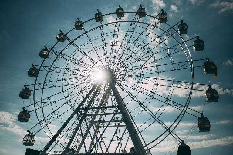 A imagem mostra uma roda gigante vista de baixo， com a estrutura metálica em destaque. O céu está parcialmente nublado， com algumas nuvens e a luz do sol visível atrás da roda. As cabines da roda gigante estão suspensas， e a perspectiva enfatiza a altura da atração