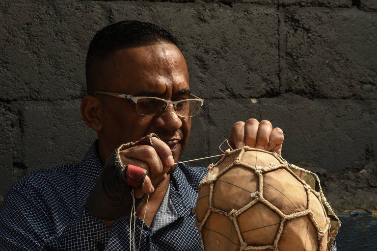 Um homem está concentrado em consertar uma bola de futebol feita de material natural. Ele usa óculos e tem cabelo curto. O fundo é uma parede de pedra cinza. O homem está segurando a bola com uma mão e costurando com a outra, utilizando um fio.