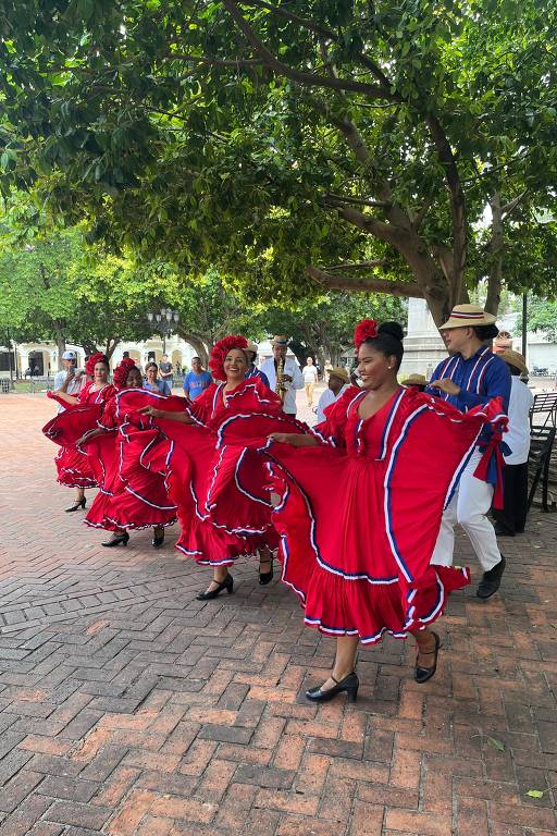 Um grupo de dançarinas vestidas com vestidos vermelhos e detalhes em azul se apresenta em uma área ao ar livre. Elas estão dançando em uma calçada de tijolos, com árvores ao fundo. Algumas pessoas assistem à apresentação, e um homem vestido de forma tradicional está ao lado delas.