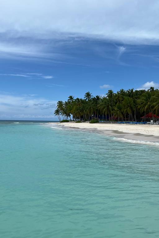 A imagem mostra uma praia tropical com águas cristalinas em tons de verde e azul. À direita, há uma faixa de areia branca e, ao fundo, uma fileira de palmeiras. O céu está parcialmente nublado, com nuvens brancas e um azul intenso.