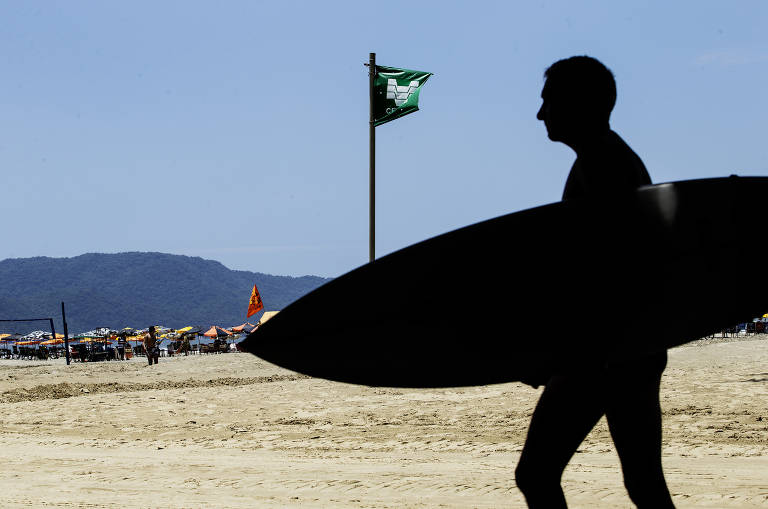 A imagem mostra a silhueta de um surfista caminhando na areia da praia, segurando uma prancha de surf. Ao fundo, há uma bandeira verde hasteada e várias pessoas sob guarda-sóis coloridos. Montanhas são visíveis ao longe sob um céu claro.