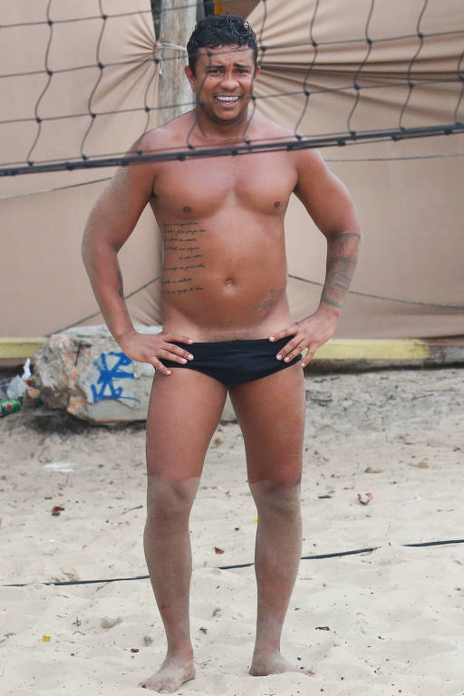 Um homem de pele morena, sorrindo, está posicionado em uma praia. Ele está sem camisa e usa um sunga preta. O fundo mostra uma rede de vôlei e uma estrutura de lona. O chão é de areia, e há algumas pedras e lixo visíveis ao fundo.