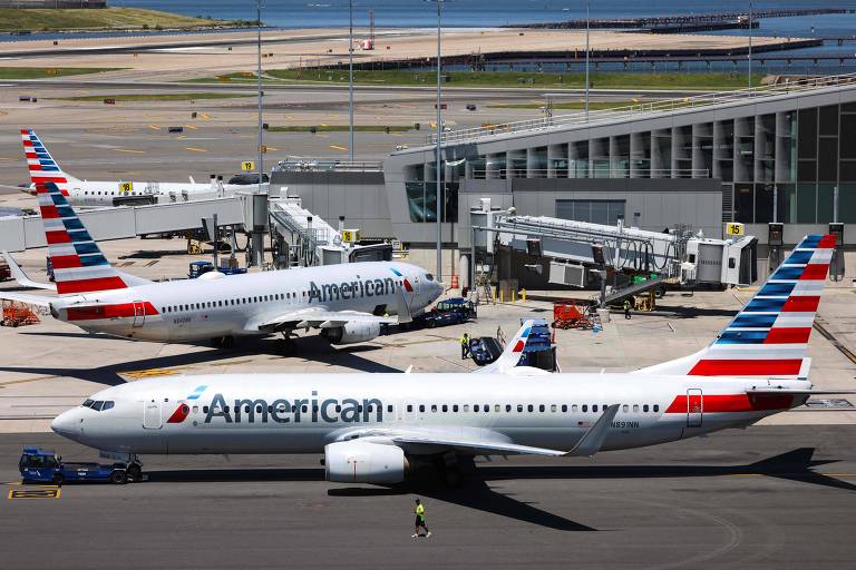 A imagem mostra vários aviões da American Airlines estacionados em um aeroporto. Um dos aviões está em primeiro plano, com a fuselagem branca e a cauda decorada com listras vermelhas, brancas e azuis. Ao fundo, há mais aviões e um terminal de aeroporto visível.