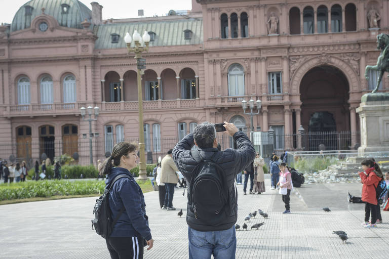 A imagem mostra um grupo de pessoas em uma praça, com um edifício histórico ao fundo. Um homem está tirando uma foto, enquanto uma mulher observa. Há outras pessoas ao redor, algumas interagindo com pombos no chão. O edifício é de cor rosa e possui janelas grandes e detalhes arquitetônicos.