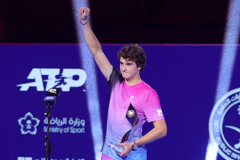 Um jovem jogador de tênis está em pé， levantando um troféu com a mão direita. Ele usa uma camiseta em tons de rosa e azul. O fundo é iluminado com luzes e apresenta logotipos de eventos de tênis. O jogador parece estar celebrando uma vitória.
