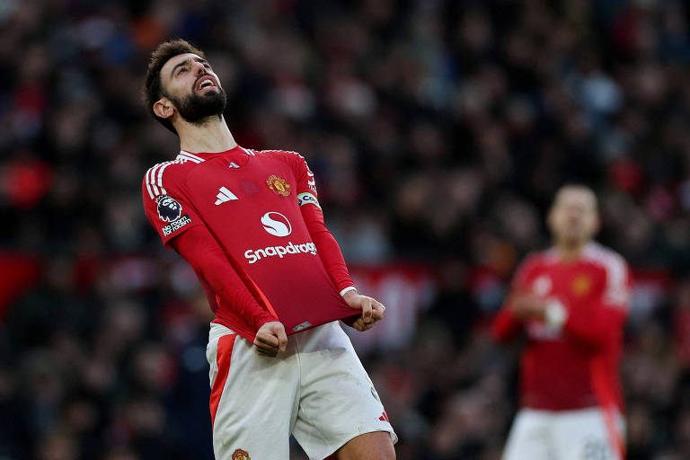 Um jogador do Manchester United， vestindo uma camisa vermelha com detalhes brancos， está em uma pose expressiva， puxando sua camisa para baixo enquanto olha para o céu. O fundo mostra uma multidão desfocada， sugerindo um ambiente de jogo intenso.