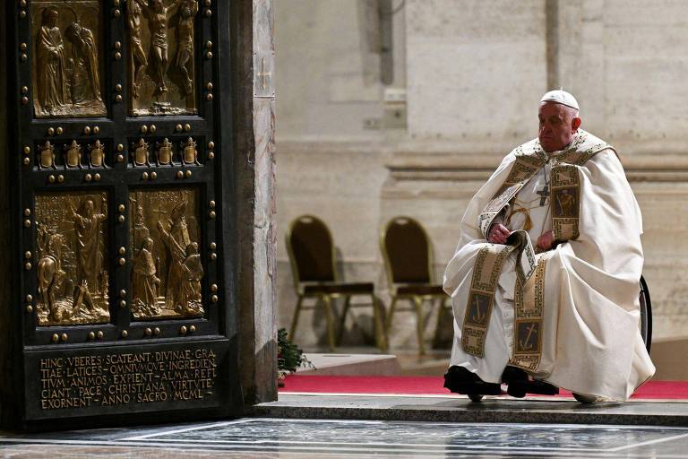 O Papa está sentado em um ambiente interno， próximo a uma porta decorada com relevos. Ele usa vestes brancas e uma estola. Ao fundo， há cadeiras vazias e um tapete vermelho no chão.