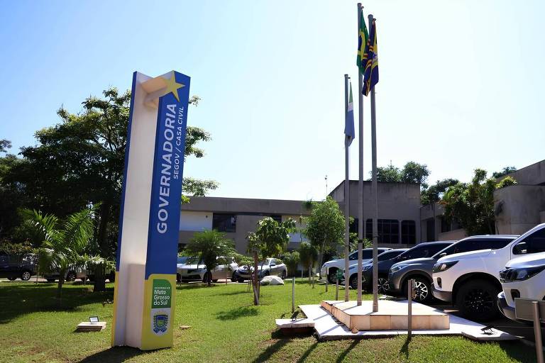 A imagem mostra a fachada de um edifício governamental com um letreiro azul que diz 039;GOVERNADORIA SEGOV / CASA CIVIL039;. Há três bandeiras hasteadas em postes ao lado do letreiro， incluindo a bandeira do Brasil. O espaço é cercado por gramado verde e algumas árvores， com vários carros estacionados em uma área visível ao fundo.