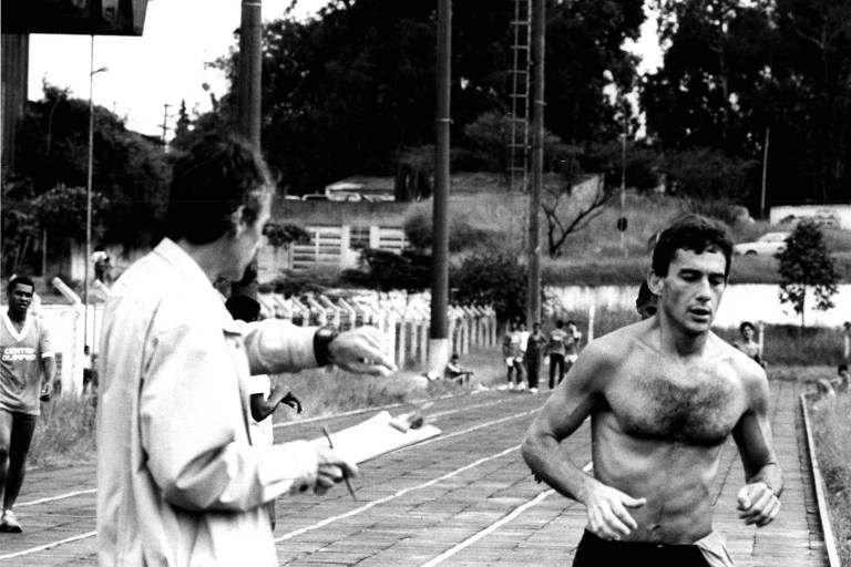 A imagem em preto e branco mostra um corredor sem camisa， com cabelo curto e uma expressão de esforço， correndo em uma pista. À sua esquerda， um homem de cabelo grisalho， vestido com um paletó claro， está segurando uma prancheta e gesticulando， possivelmente dando instruções ou anotando algo. Ao fundo， há outros corredores e uma área verde.