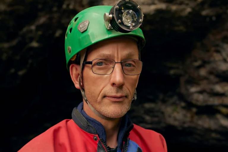 Um homem com um capacete verde equipado com uma lanterna frontal， usando óculos e uma roupa de proteção vermelha. Ele está posicionado em um ambiente rochoso， com uma expressão séria， olhando diretamente para a câmera