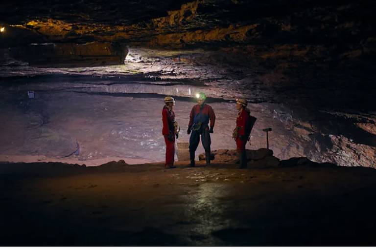 A imagem mostra três pessoas em uma mina subterrânea. Eles estão vestidos com roupas de proteção vermelhas e capacetes com lanternas. O ambiente é escuro， com iluminação suave proveniente de luzes no teto da mina. O chão é de terra e rocha， e as paredes da mina são irregulares e de cor marrom. As pessoas estão em pé， conversando entre si