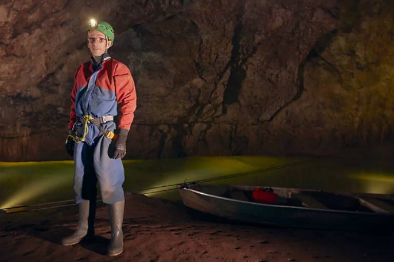 Um homem está posicionado em uma caverna， vestindo um macacão azul e uma jaqueta vermelha. Ele usa um capacete com uma lanterna na frente e luvas. Ao fundo， há uma superfície de água iluminada em tons de verde， e um pequeno barco à direita， ancorado na areia