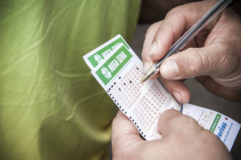 A imagem mostra uma pessoa segurando um bilhete de loteria da Mega-Sena， enquanto escreve com uma caneta. O bilhete é branco com detalhes em verde e contém números marcados. A pessoa está vestindo uma camisa amarela.