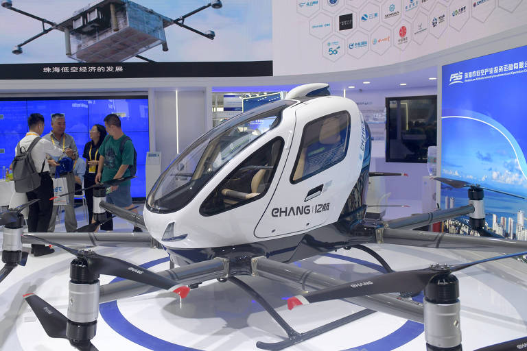 A imagem mostra um veículo aéreo elétrico em uma exposição. O veículo, com design futurista, possui uma fuselagem branca e é apoiado em quatro hélices. Ao fundo, há um painel com uma imagem do veículo em voo e várias pessoas observando e interagindo com a exibição. O ambiente é moderno, com iluminação e elementos gráficos nas paredes.