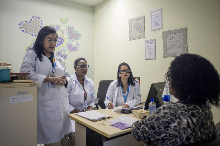 A imagem mostra um ambiente de consulta médica， onde três profissionais de saúde， vestindo jalecos brancos， estão em pé e observando uma paciente sentada à mesa. As profissionais têm expressões sérias e estão atentas à conversa. A paciente， que está de costas， parece estar respondendo a perguntas. O ambiente é decorado com desenhos de corações e há documentos e um computador sobre a mesa. Na parede， há quadros com textos motivacionais.