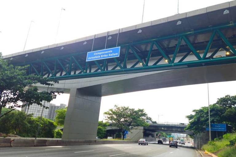 ponte recém construída, com placa azul fixada em sua estrutura, cruza sobre avenida