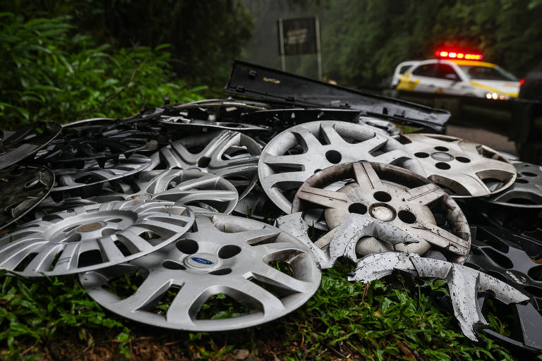 A imagem mostra um monte de calotas de carro, algumas em bom estado e outras danificadas, empilhadas em um terreno coberto de grama. Ao fundo, é possível ver um veículo de polícia com luzes de emergência acesas, em um ambiente de floresta densa e nevoeiro.