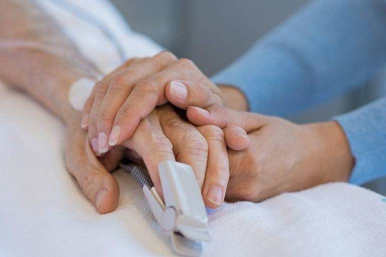 Foto mostra a mão esquerda de um paciente branco sendo segurada pelas duas mãos de um profissional de saúde também branco. A mão do paciente tem um aparelho no dedo que serve para medir sinais vitais. 