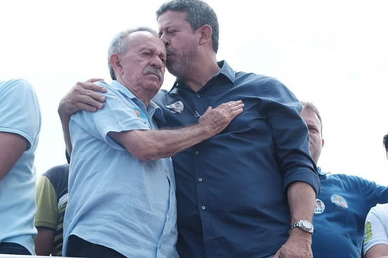 Foto mostra Benedito (à esq.) recebendo um abraço e um beijo de Arthur (à dir.). Os dois são homens de pele clara. Arthur é maior que o pai e veste uma camisa azul escura. Ele tem cabelos curtos e grisalhos. Benedito usa uma camisa azul clara e tem cabelos grisalhos com sinais de calvície. Ele também ostenta um bigode da mesma cor que o cabelo.